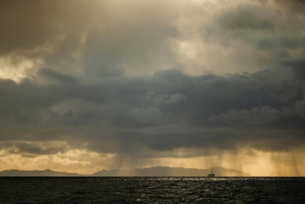 Dramatische Meereslandschaft mit dunklen Regenwolken am Himmel. Sonnenstrahlen durchbrechen die Wolkendecke und beleuchten die Silhouette einer Insel am Horizont. Ein Schiff fährt in der Ferne über das Wasser.